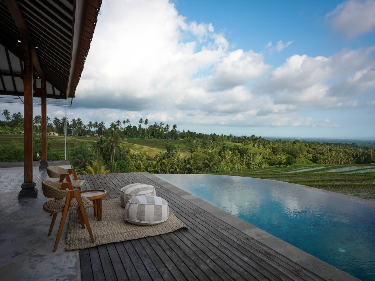 An outdoor space features an infinity pool overlooking lush green rice terraces and palm trees. Natural wood decking surrounds the pool, complemented by two woven chairs and large floor cushions, providing a comfortable area for relaxation against a serene backdrop of the countryside.