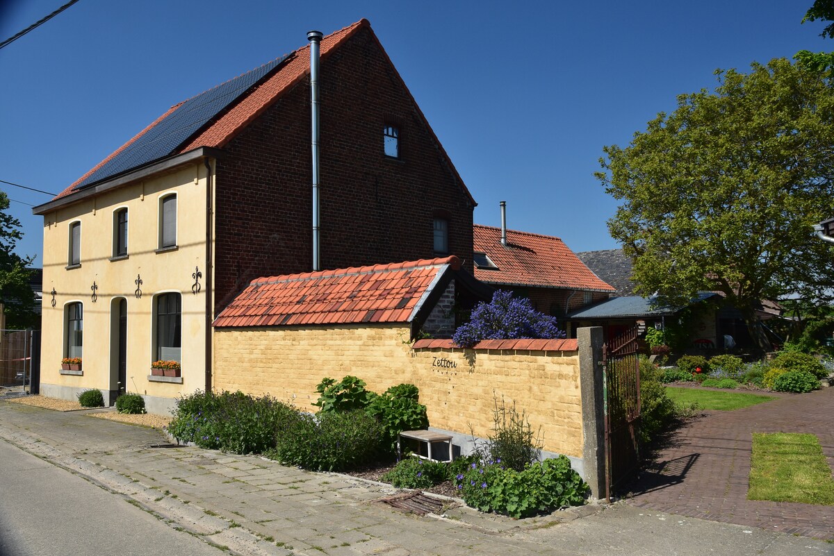 The charming exterior of a two-story house is presented, featuring a combination of yellow plaster and red brick. Flower boxes are visible under the windows, and a well-maintained garden can be seen in the front yard, enhancing the welcoming appearance.