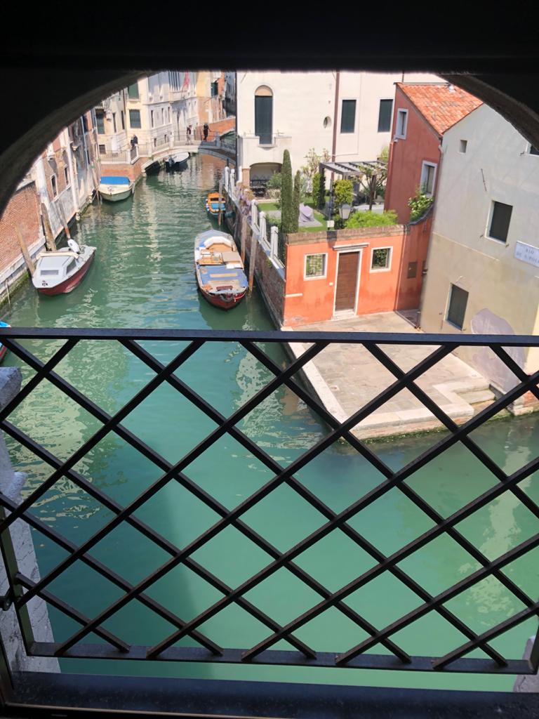 A view through a decorative wrought-iron balcony showcases the serene canals of Venice. Colorful buildings line the water, reflecting shades of green and blue. Several boats are moored at the edges, while a quaint pathway leads to a charming garden in the background.