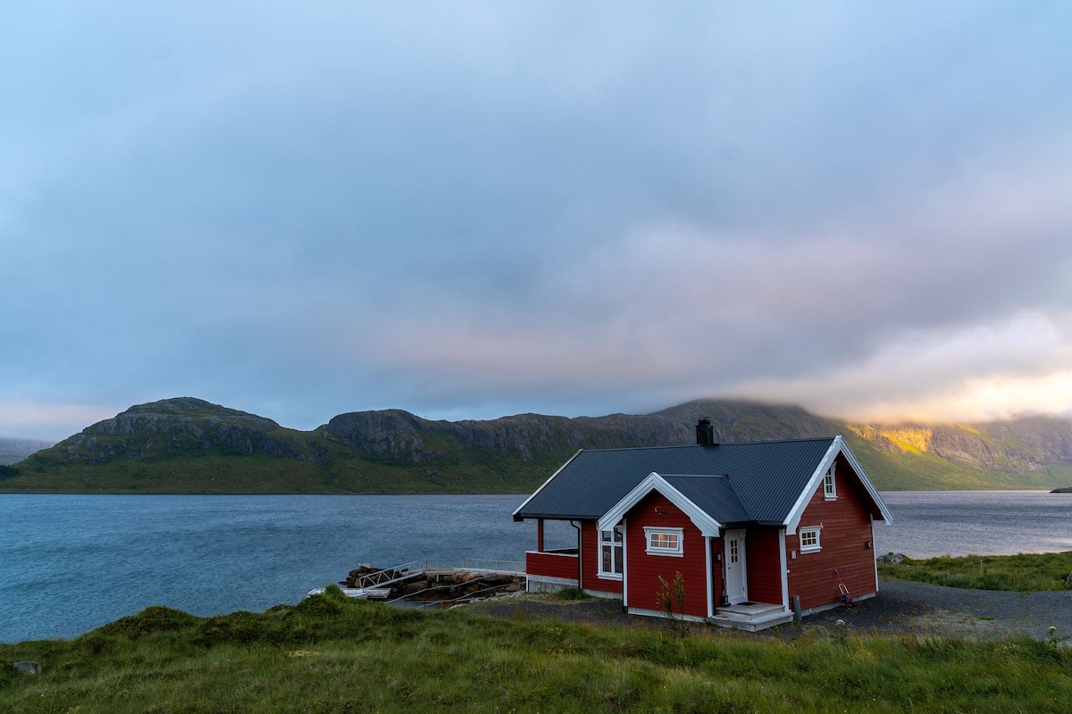 A red cabin is situated by a tranquil body of water, surrounded by green hills. The structure features a black roof and white accents, with windows highlighting the beautiful natural scenery. Soft clouds hover above, creating a serene atmosphere.