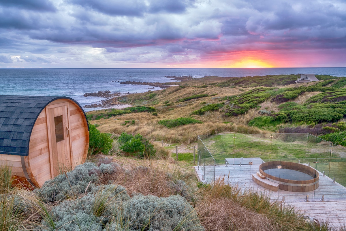A wooden sauna is positioned near the cliff's edge, offering views of the ocean and coastline. A circular hot tub sits adjacent on the deck, surrounded by lush coastal vegetation. The sky above displays a colorful sunset, reflecting hues of orange and pink across the water.