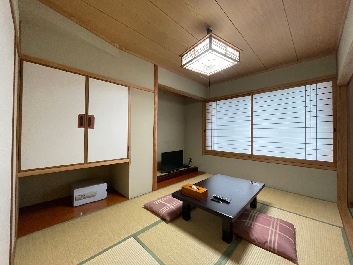 A traditional Japanese room features tatami flooring and a low wooden table with floor cushions. A television is positioned on a shelf, and a wall cabinet is visible. Natural light filters through window slats, creating a serene ambiance.