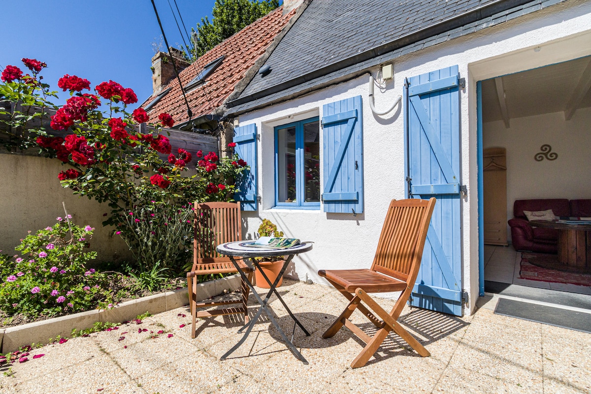 A small outdoor patio space features two wooden foldable chairs beside a quaint table, set against a backdrop of vibrant red roses and blue shutters. Sunlight illuminates the area, creating a welcoming environment for relaxation.