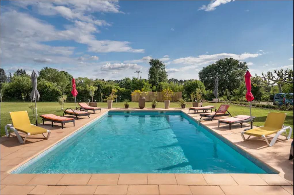 A swimming pool is situated at the center of a spacious outdoor area, surrounded by lounge chairs and umbrellas. Lush greenery and trees are visible in the background, enhancing the serene atmosphere. The pool features a clear blue water surface reflecting the sky.