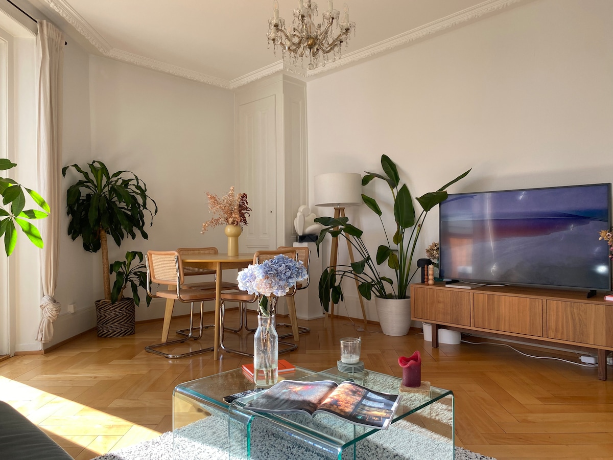 A bright living area features a glass coffee table with a magazine and a small candle. Lush green plants surround the space, including a large potted plant near a wooden cabinet with a television. A dining table with chairs is positioned in the background, under soft natural light.