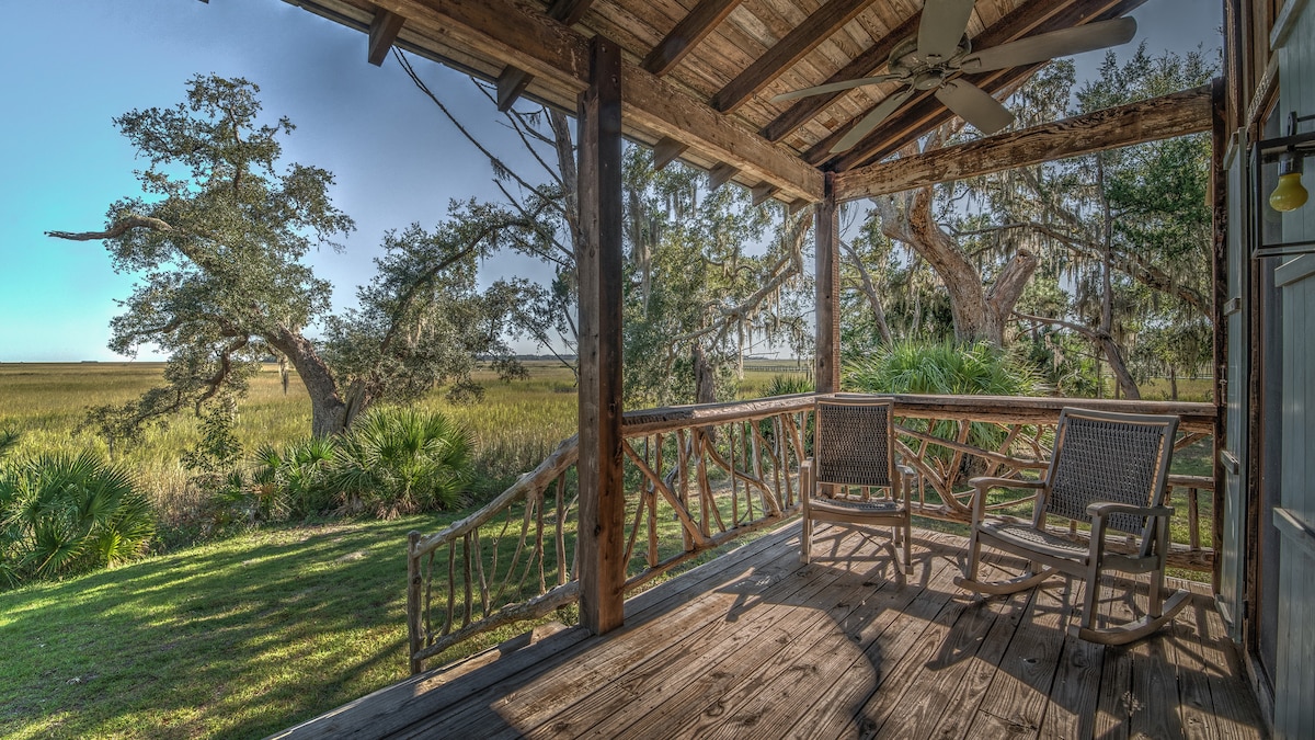 A serene porch area features wooden flooring and a rustic railing, offering a welcoming spot to relax. Two comfortable chairs are positioned for enjoying views of the marsh and live oaks, with sunlight filtering through the foliage.