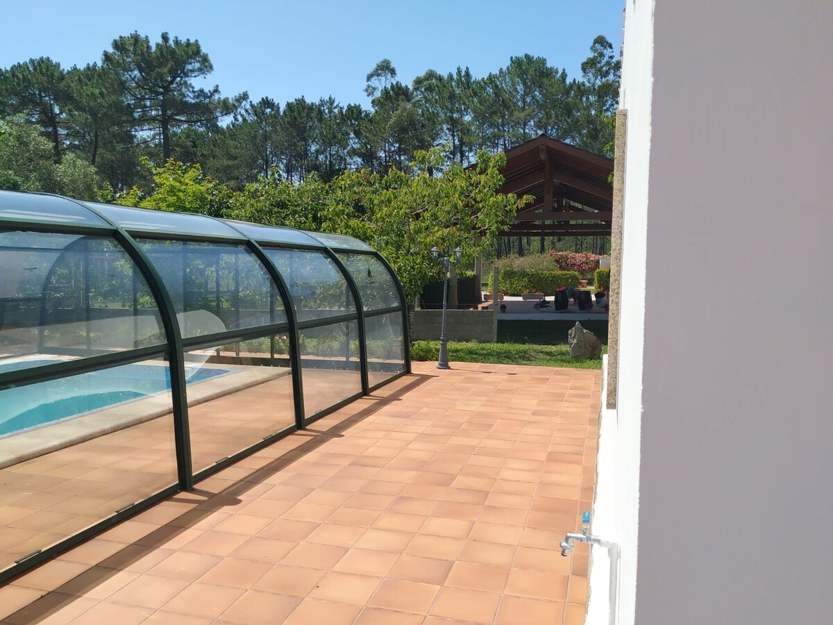 A pool area is enclosed with glass, allowing natural light to illuminate the space. A spacious terrace features tiled flooring, with greenery and trees visible in the background. A large structure with a wooden roof is seen beyond the pool area, suggesting relaxation and nature.