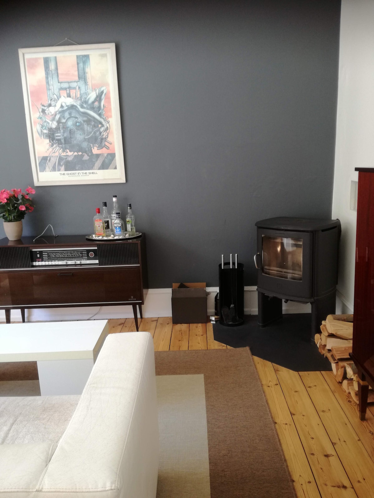 A welcoming living area features a neutral-colored wall with a distinctive art piece above a stylish sideboard. A compact wood-burning stove is positioned nearby, with a stack of logs beside it. A plush white sofa and a light area rug complete the space.
