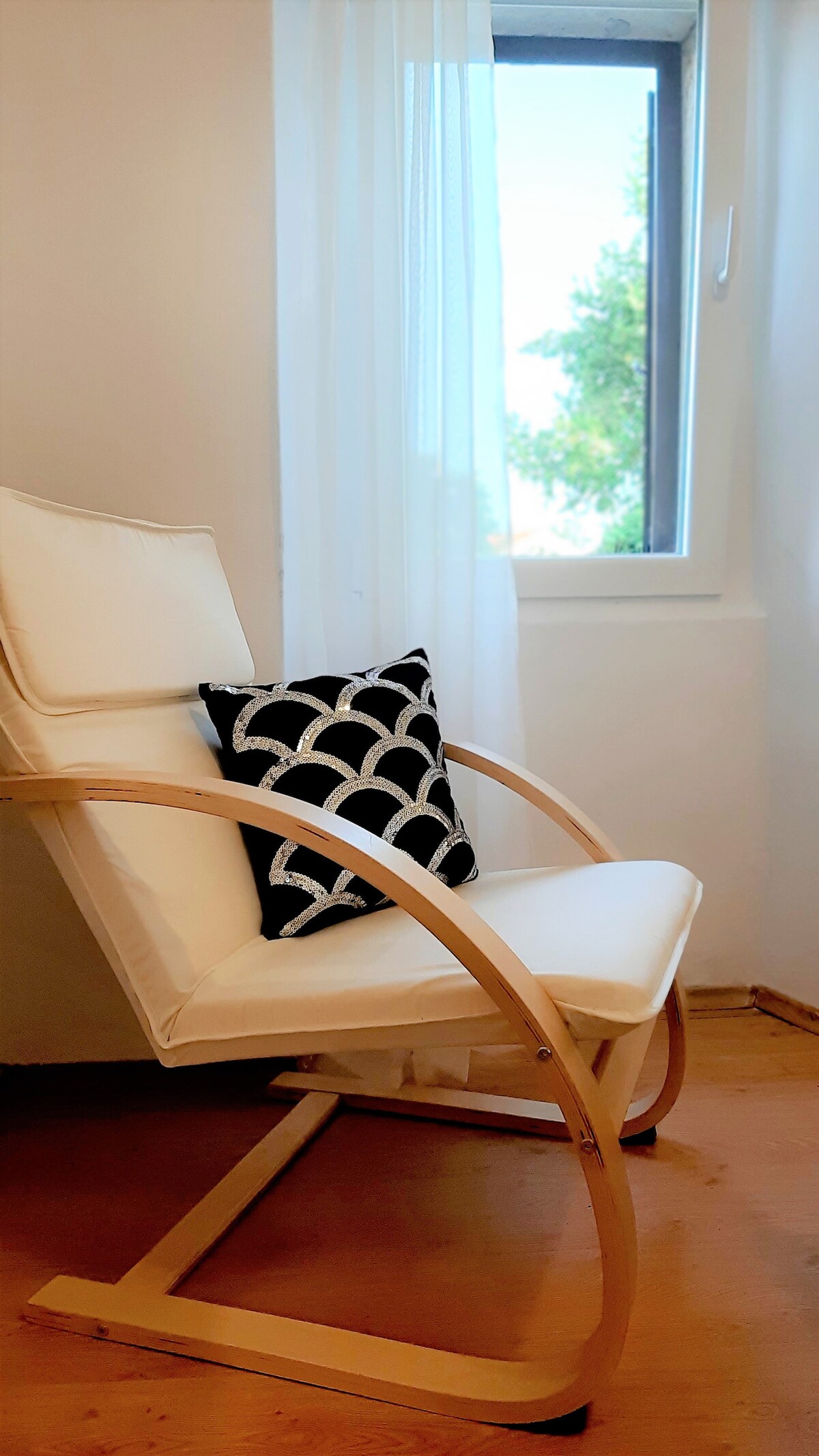 A modern rocking chair is positioned near a window, allowing natural light to enter the space. A decorative black and white pillow adds a touch of style to the chair. Sheer curtains frame the window, creating a soft ambiance.