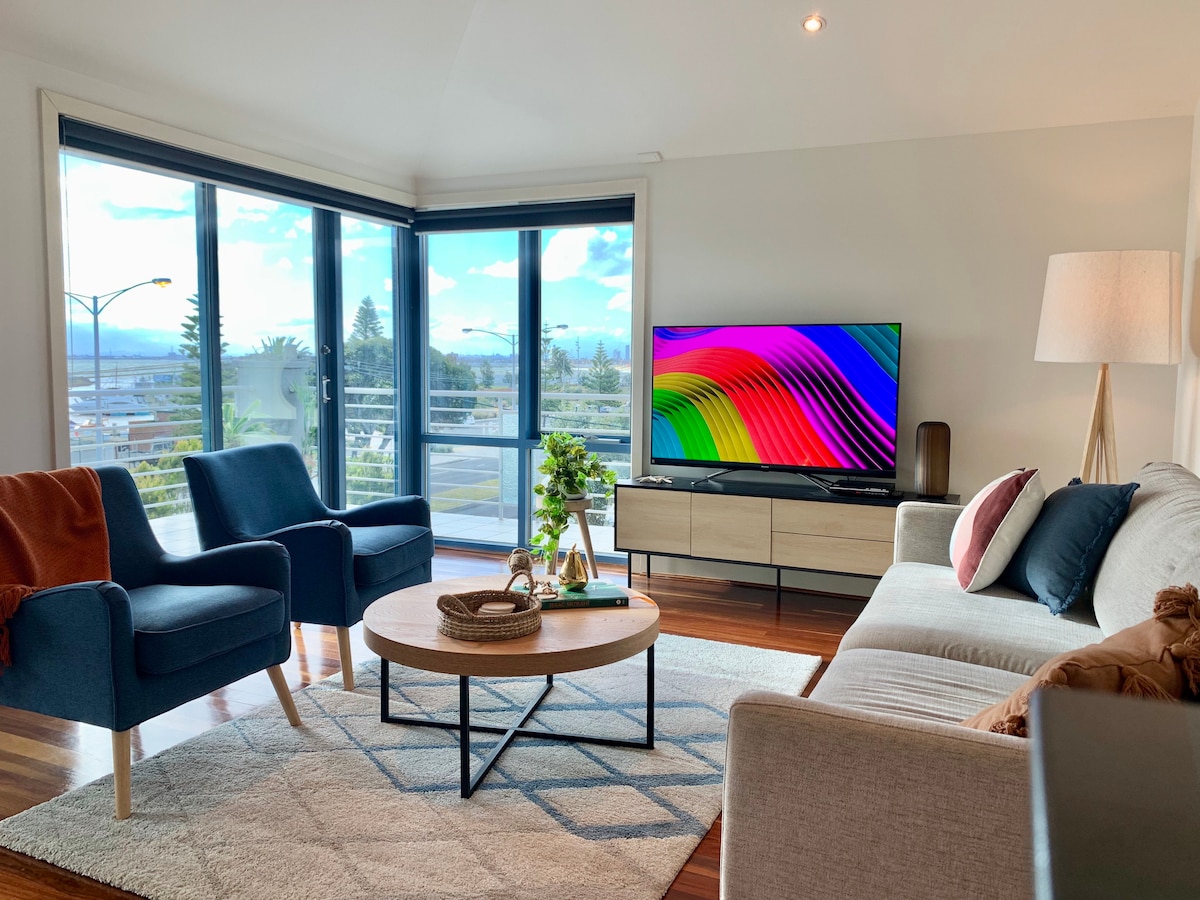 An open-plan living area is showcased with a spacious sofa and two accent chairs, surrounding a round coffee table. A large flat-panel TV displays colorful imagery, while floor-to-ceiling windows provide views of the outdoors and allow natural light to fill the room.