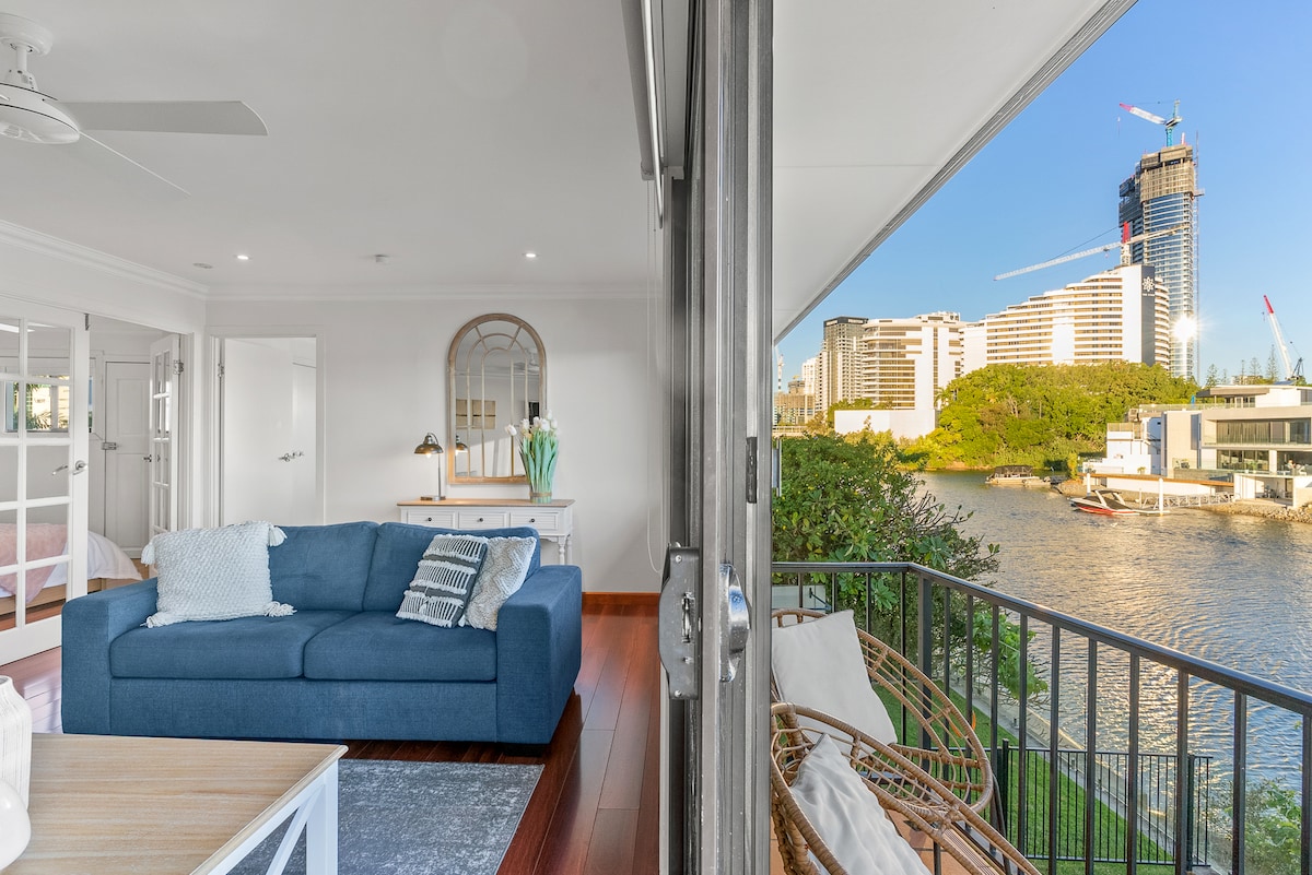 A comfortable living area features a blue couch with light cushions, complemented by a nearby console table and decorative mirror. Large glass doors open to a balcony overlooking the waterfront, with modern buildings and boats visible in the background.