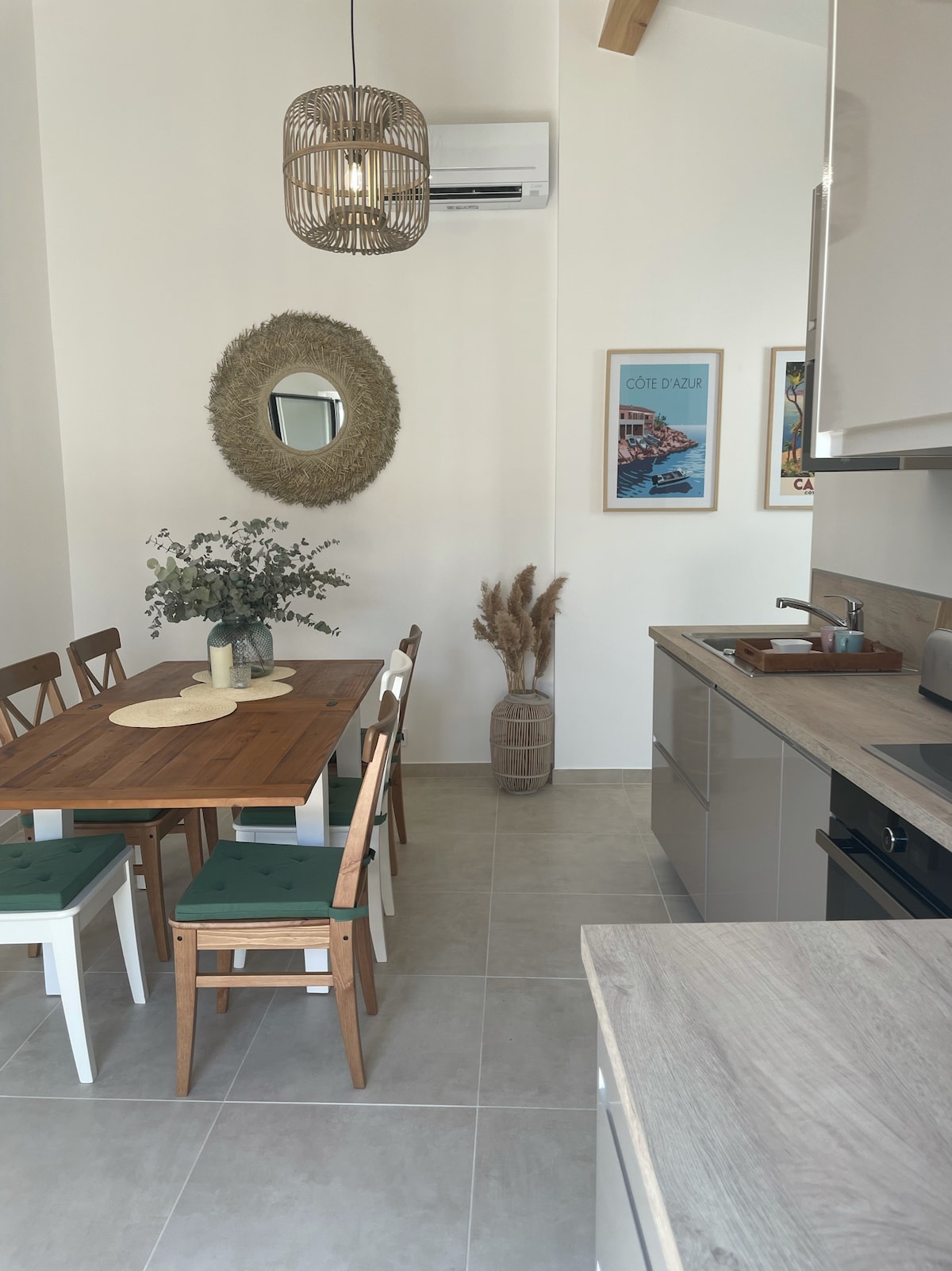A dining area features a wooden table surrounded by six chairs with green cushions. A round mirror with a natural weave hangs on the wall, while a vase with dried plants adds a touch of decor. The space enjoys a bright, open layout.