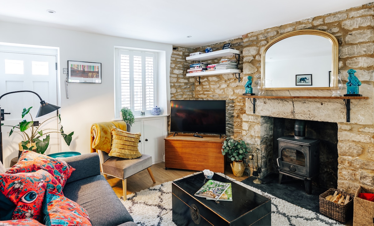A cozy living room features a stone fireplace with a log burner, enhancing the space. A Smart TV is positioned on a wooden cabinet. A comfortable sofa, adorned with vibrant cushions, complements a bright armchair. Shelves display books and decorative items, while plants add a natural touch.