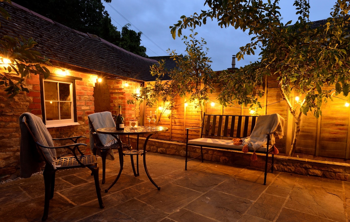 A private courtyard garden is illuminated by soft string lights, creating a warm ambiance. A black metal table is set with two glasses, accompanied by a comfortable bench draped with cozy blankets. Lush greenery enhances the peaceful outdoor space.