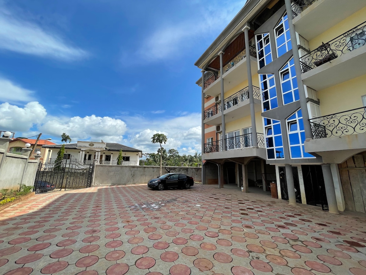 An expansive parking area is visible in front of a three-story building, which is adorned with decorative balconies. The ground is paved with circular red and cream tiles. A black car is parked, and greenery and neighboring buildings are seen in the background under a clear blue sky.