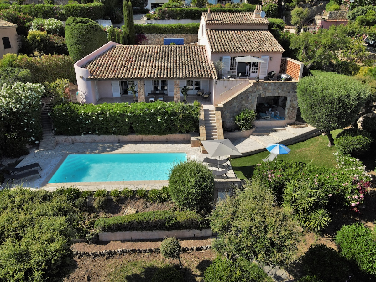 An aerial view of a villa surrounded by lush greenery showcases a tranquil swimming pool in the foreground. A spacious patio with chairs is visible, and the main house features a terracotta roof and large windows, inviting natural light into the living areas.