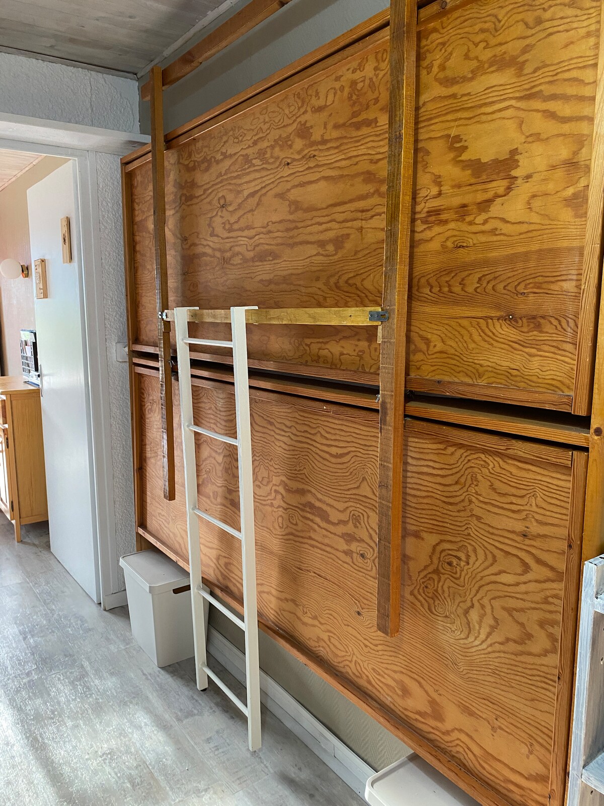 A wooden wall features a mounted fold-down bunk bed, with a white ladder for access. Two beige storage bins are placed on the floor beneath the structure, offering convenient organization. The natural wood grain adds texture to the space.