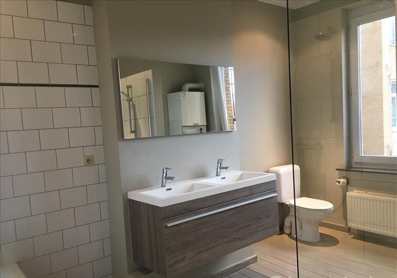 A modern bathroom is showcased, featuring a dual sink with a wooden vanity. A large mirror reflects the well-lit space. A window allows natural light while providing a view of the outside. Wall tiles in light colors complement the overall design, promoting a clean aesthetic.
