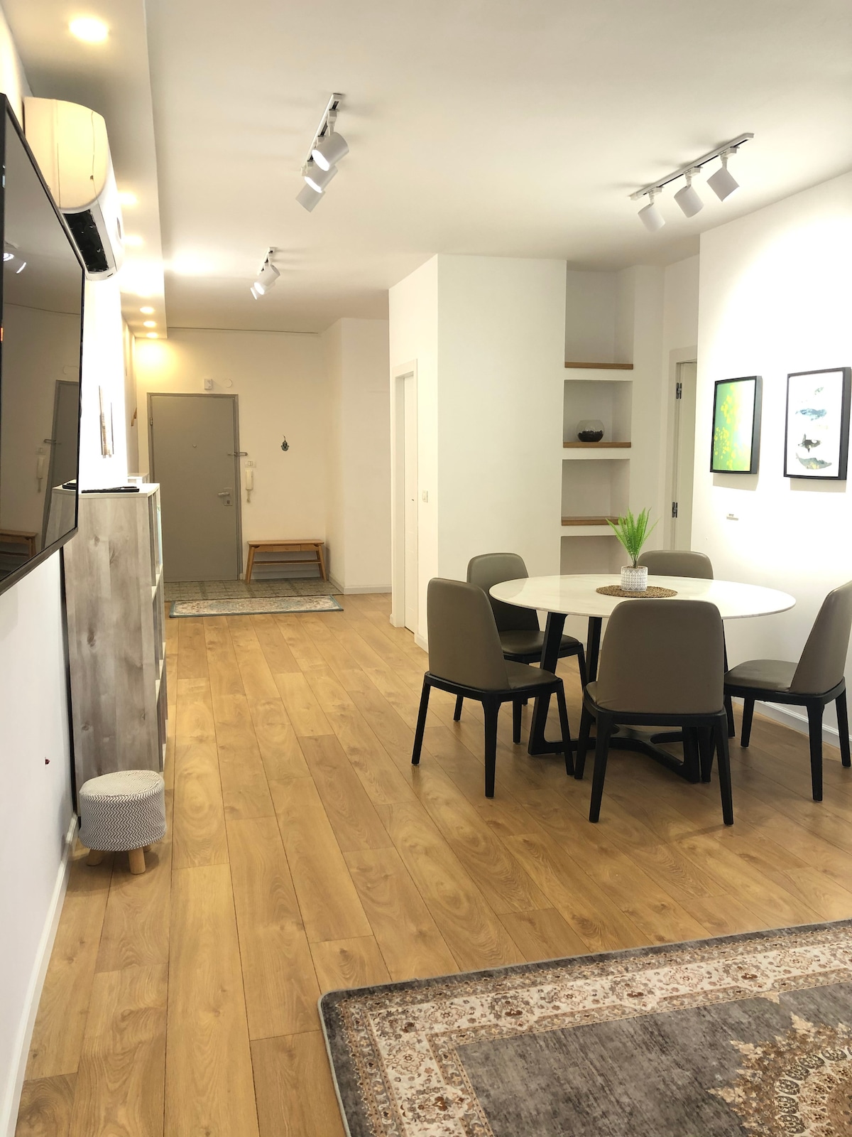 A modern dining area features a round table surrounded by four upholstered chairs. Soft lighting from recessed fixtures illuminates the space, while a small decorative plant is placed at the center of the table. Light wooden flooring extends throughout the area, adding warmth to the ambiance.