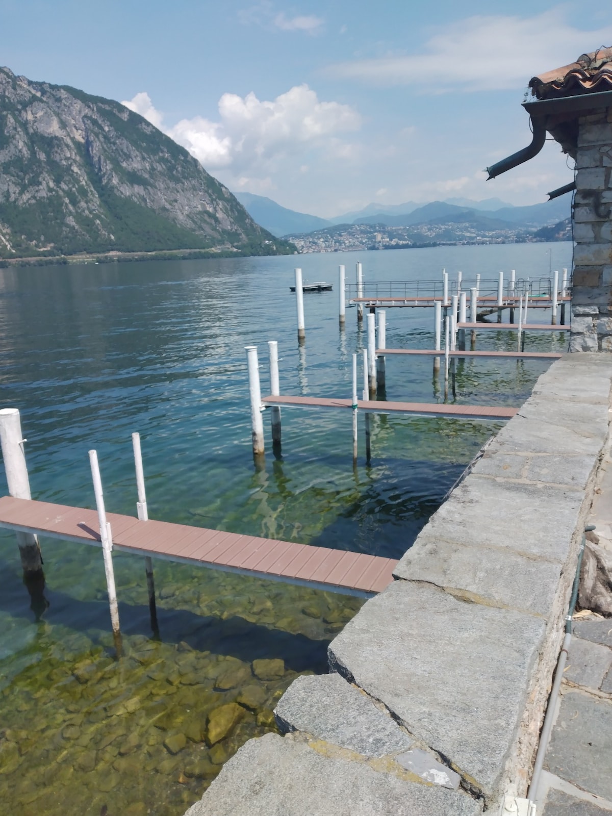 A serene lakeside view is presented, featuring wooden piers extending into the clear water. The shoreline is lined with smooth stones, while distant mountains and a blue sky create a peaceful backdrop.