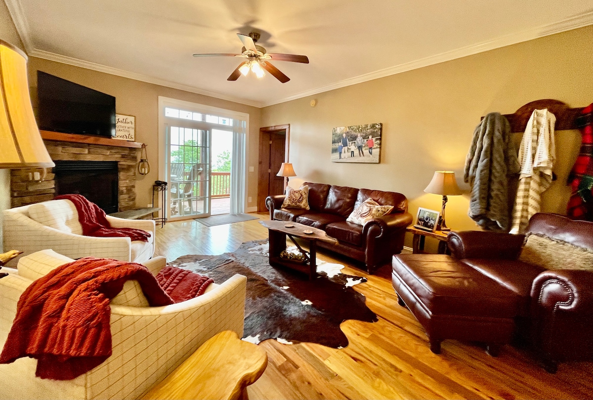 A cozy living room features a brown leather sofa, a matching armchair, and two light-colored chairs with soft blankets draped over them. A wooden coffee table sits atop a decorative rug. Natural light fills the space, highlighting the stone fireplace and the balcony visible through sliding glass doors.