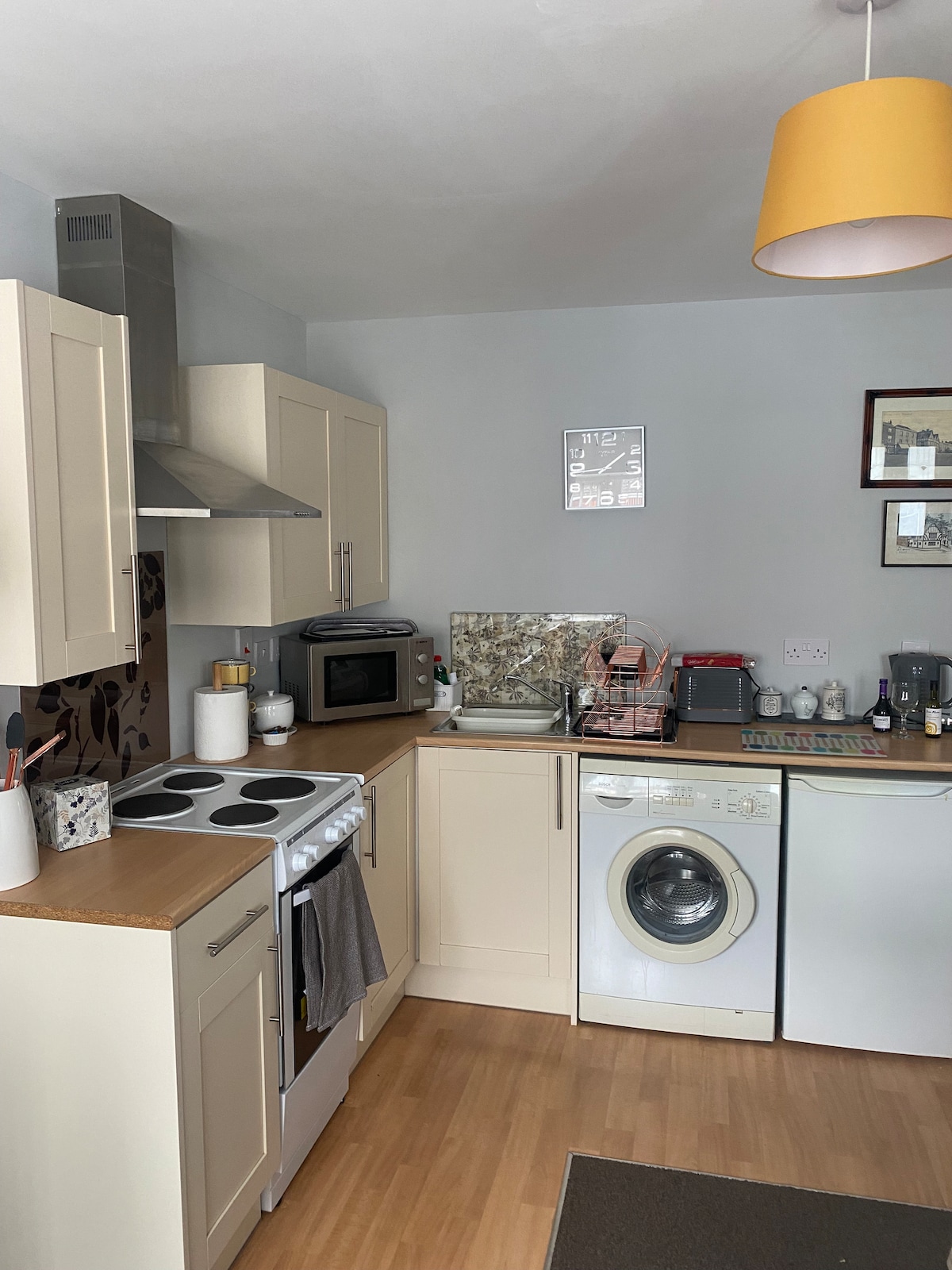 A compact kitchen space includes modern cabinetry and essential appliances, such as a stove, microwave, and washing machine. Countertop items, like dishware and utensils, are organized for convenience. A warm light fixture enhances the room's functionality and comfort.