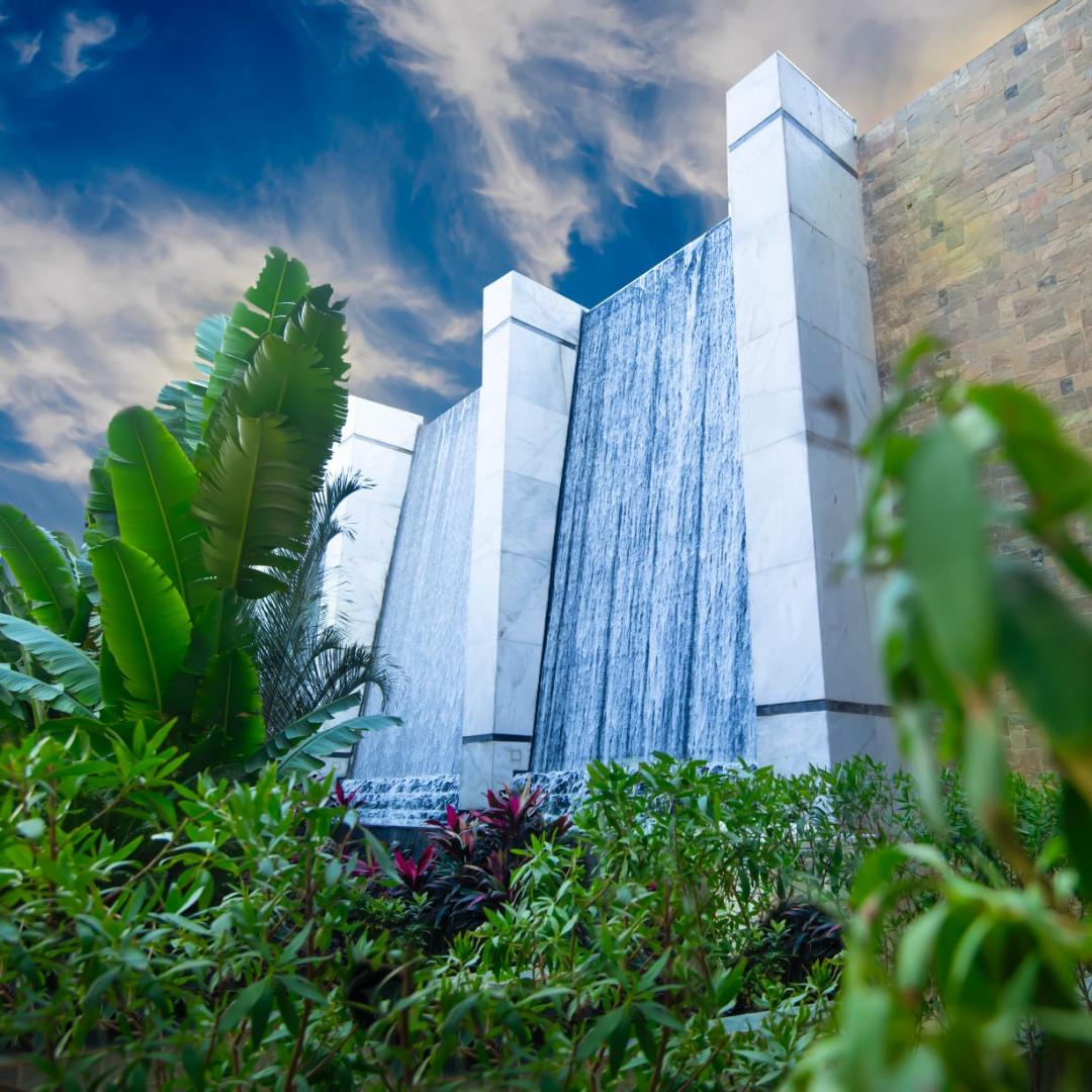 A serene waterfall feature cascades down a marble backdrop, surrounded by lush greenery. Tropical plants in the foreground create a vibrant contrast against the smooth stone walls, while a clear blue sky with wispy clouds serves as an inviting backdrop.