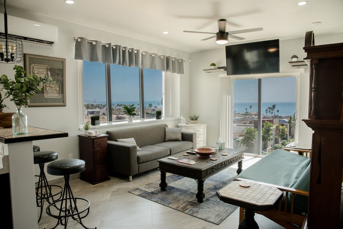 A bright living area is furnished with a gray sofa, a wooden coffee table, and a side cabinet. Large windows frame ocean views and allow natural light to fill the space. Bar stools are positioned near the kitchen counter, enhancing the open layout.