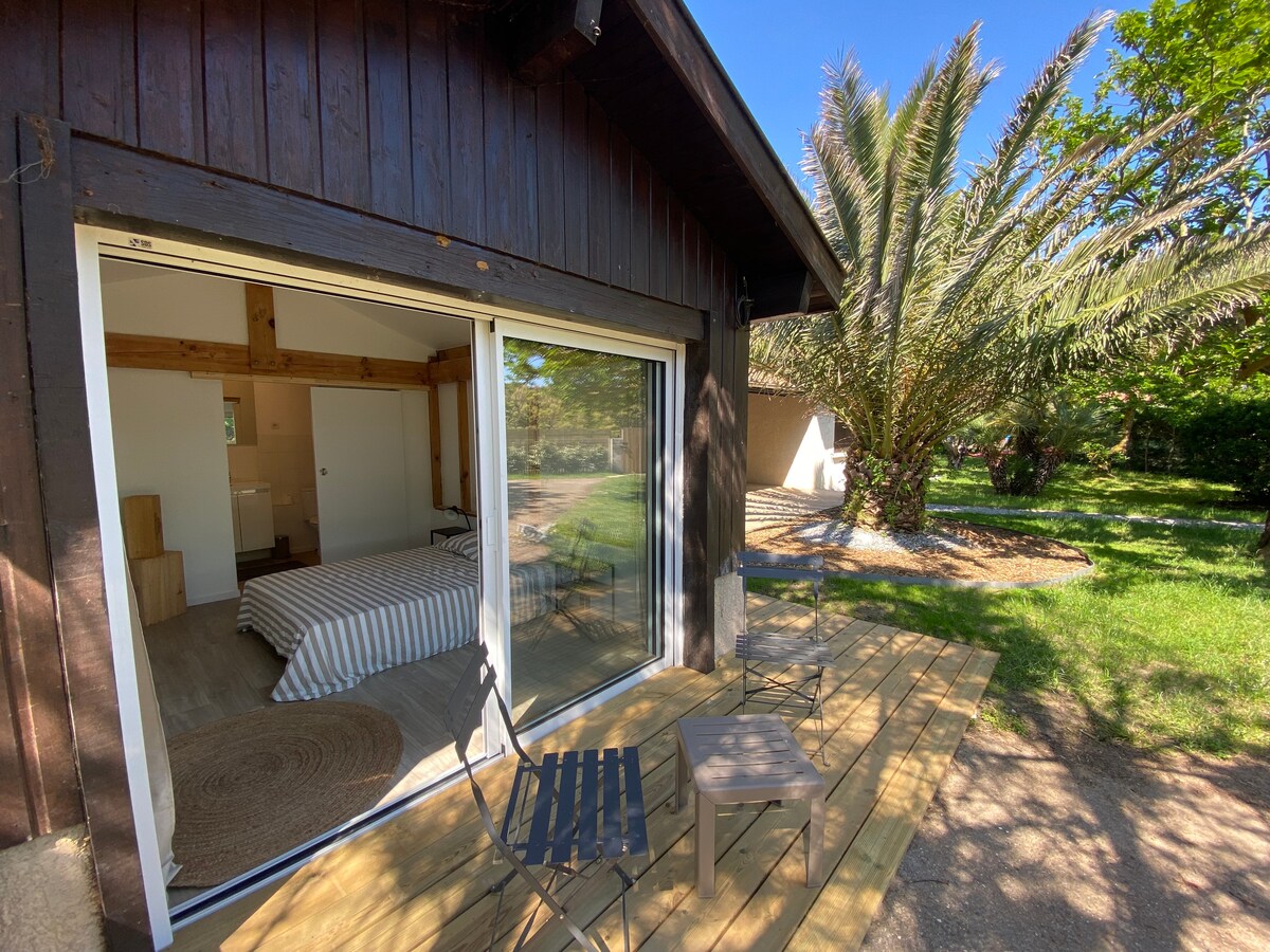 A compact exterior view of a room features large glass sliding doors opening to a wooden deck. The area is surrounded by lush greenery, including palm trees. A striped bed is visible inside, complemented by a small outdoor seating arrangement of two chairs.