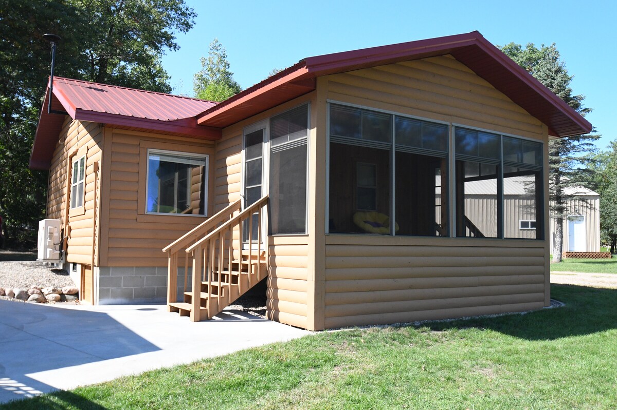 A rustic cabin features a screened-in porch and a welcoming exterior with wooden siding and a red roof. A gentle slope leads to the entrance, and the surrounding landscape includes a well-maintained lawn and gravel path, enhancing the outdoor appeal.
