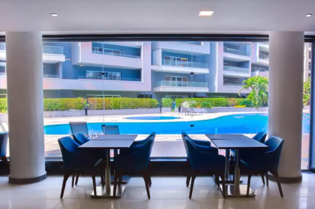 A spacious dining area is featured with dark wood tables and blue upholstered chairs arranged to face large glass windows. Outside, a swimming pool and landscaped gardens are visible, providing a view of the vibrant outdoor environment.