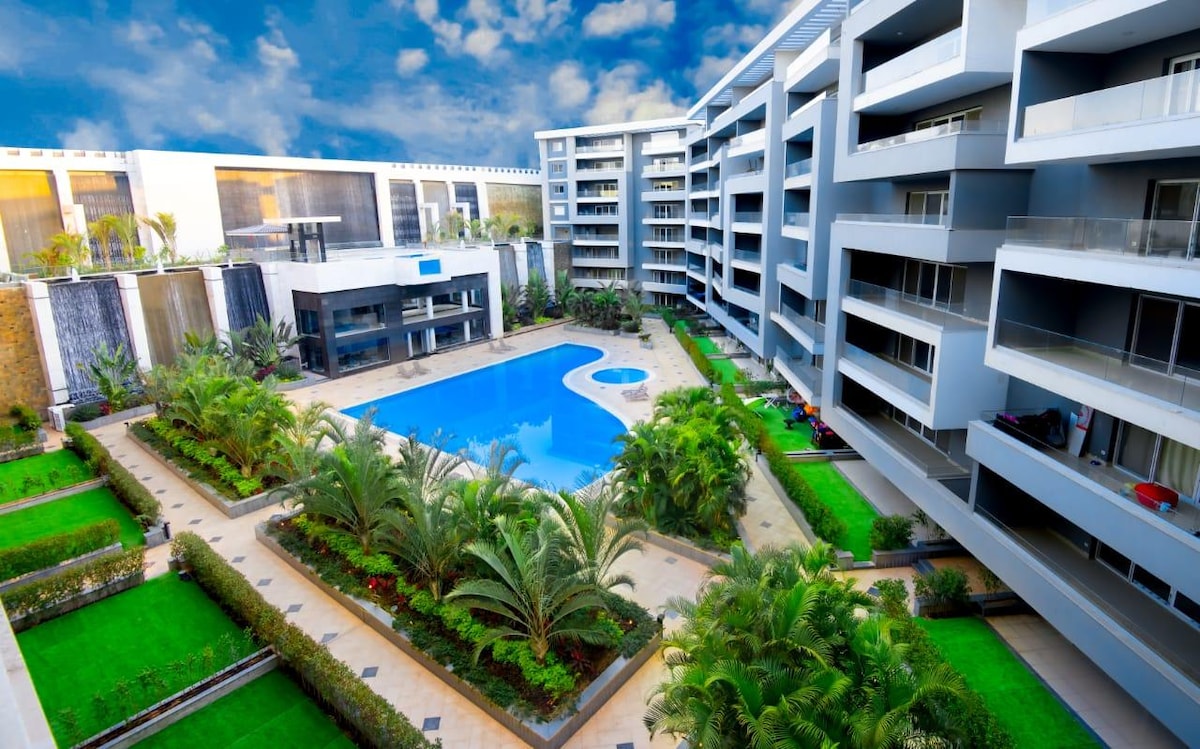 An expansive swimming pool is set in the center of a landscaped courtyard, surrounded by lush palm trees and manicured gardens. Modern buildings frame the space, with balconies overlooking the pool area, enhancing the inviting atmosphere of the community.
