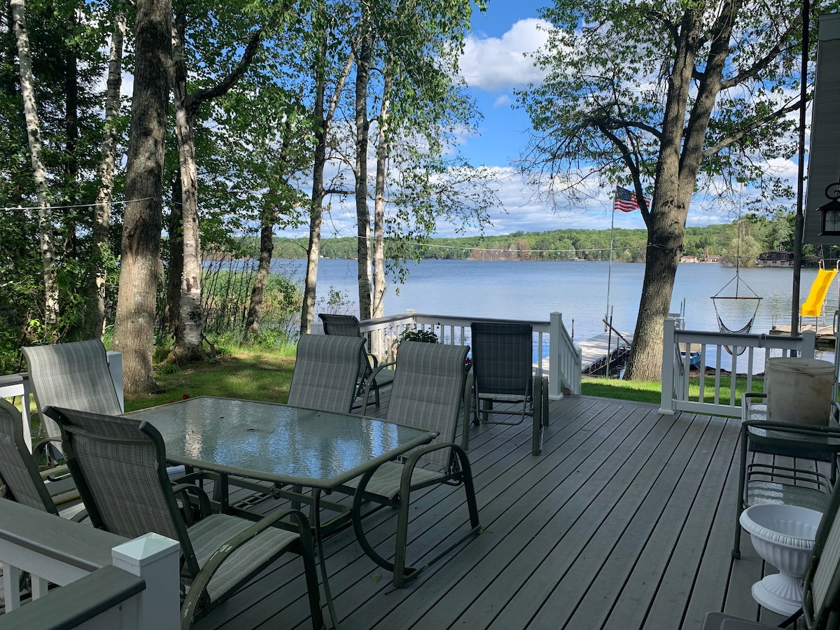 A spacious deck overlooks Archibald Lake, framed by tall trees. Multiple comfortable chairs surround a glass table, inviting relaxation. In the background, the tranquil water reflects the sky and greenery, while recreational amenities are visible at the lakeside.