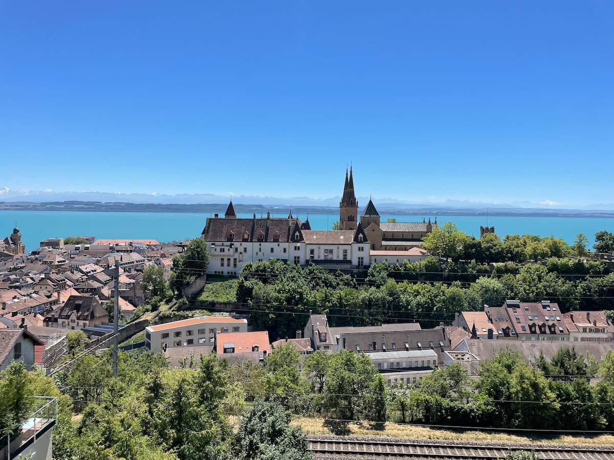 A panoramic view captures a historic building nestled among lush greenery, facing a tranquil lake under a clear blue sky. The rooftops of nearby structures are visible, with distant mountains framing the horizon.