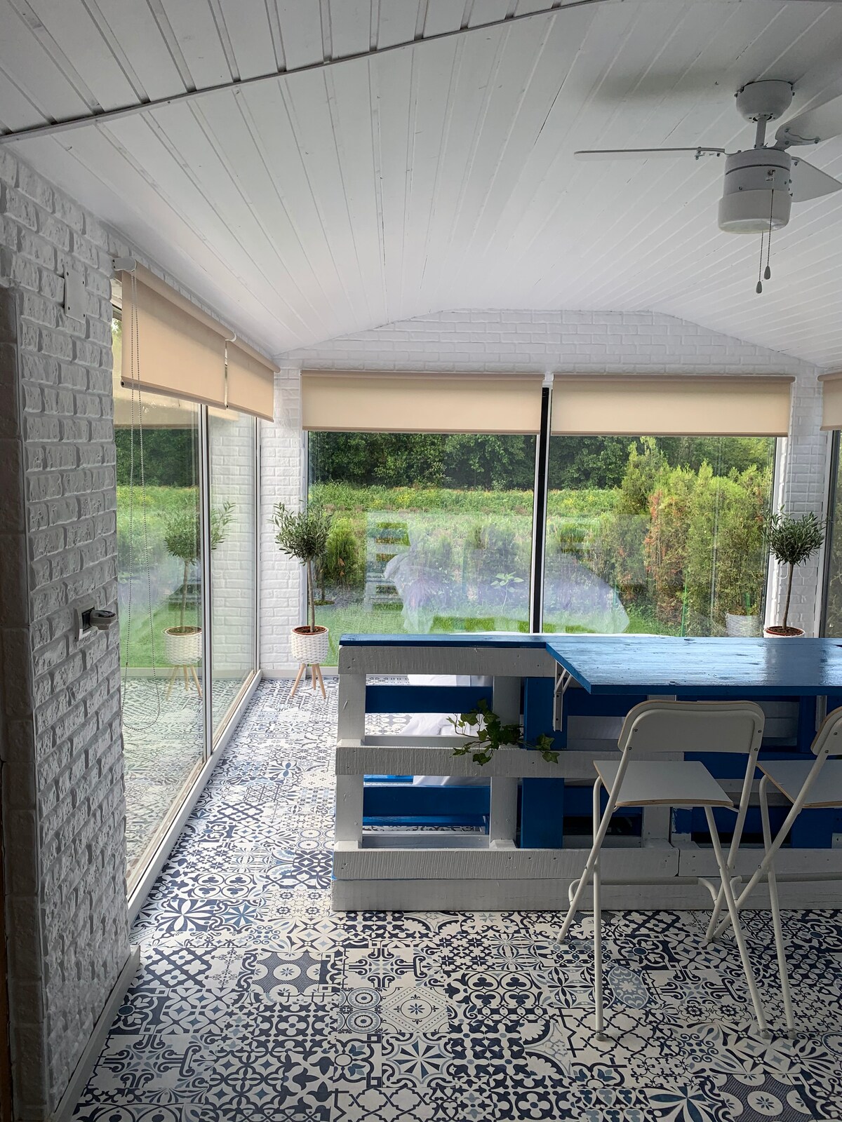 A sunlit dining area features a blue table accompanied by white chairs. Large glass doors allow views of the greenery outside, while patterned tiles add a touch of design. Soft natural light illuminates the space, creating an inviting atmosphere.