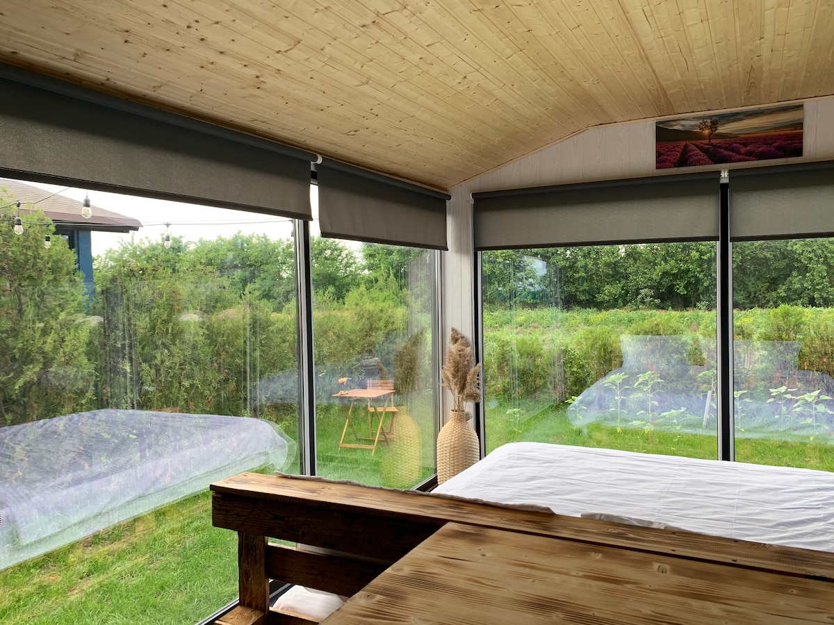 A spacious area features large windows that provide views of the lush greenery outside. Natural light floods the room, highlighting a wooden table and a neatly made bed. The wooden ceiling adds warmth, while soft lighting fixtures are positioned above.