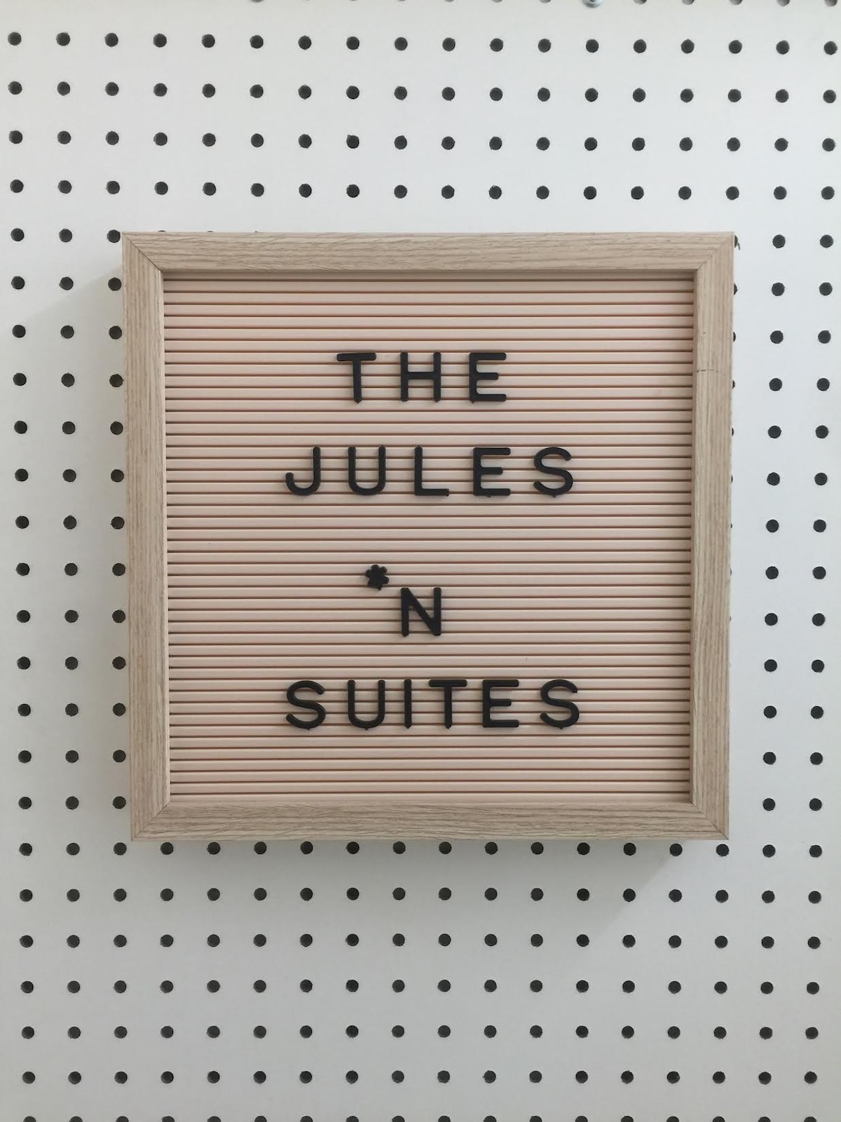 A decorative sign is displayed within a simple wooden frame. The sign features the words 'THE JULES *N SUITES' arranged in black letters on a soft pink background, positioned against a backdrop of clean, white perforated panels.