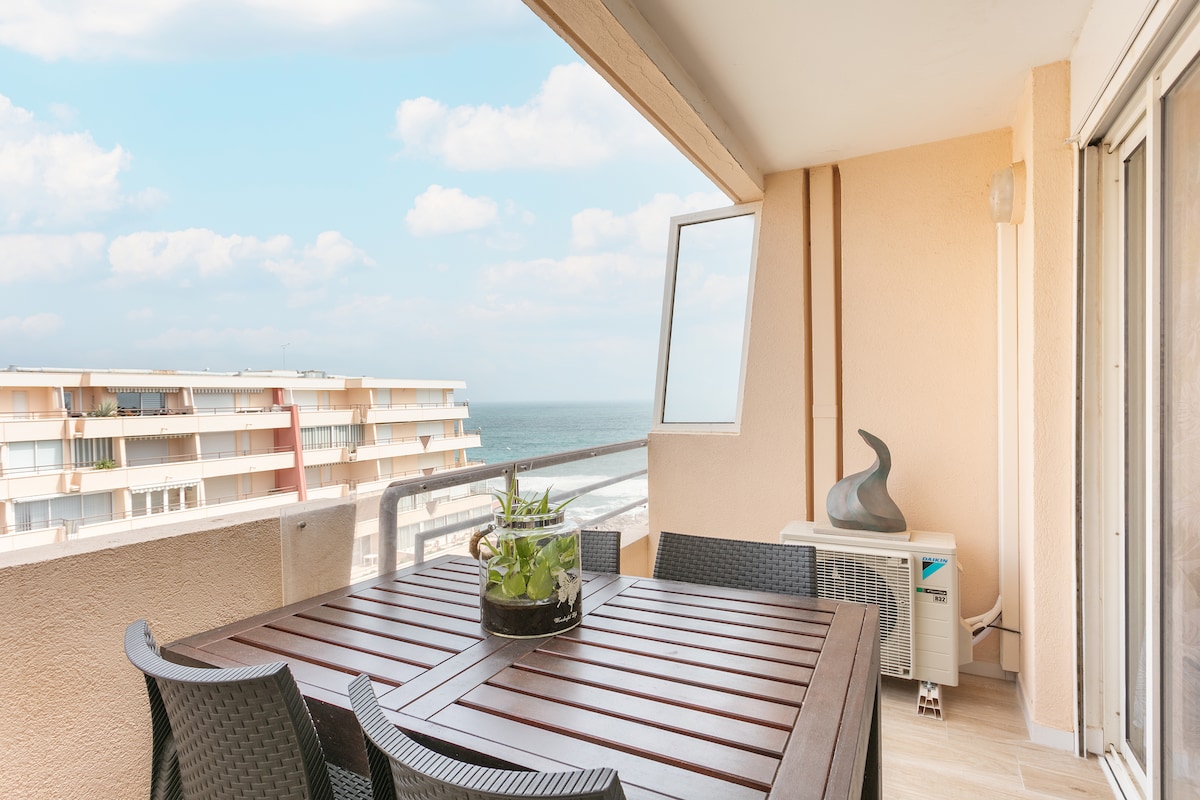 A balcony features a wooden table surrounded by six dark wicker chairs, with a potted plant centerpiece. An air conditioning unit is visible to the side, and a view of the distant sea and coastal buildings is provided through large glass sliding doors.