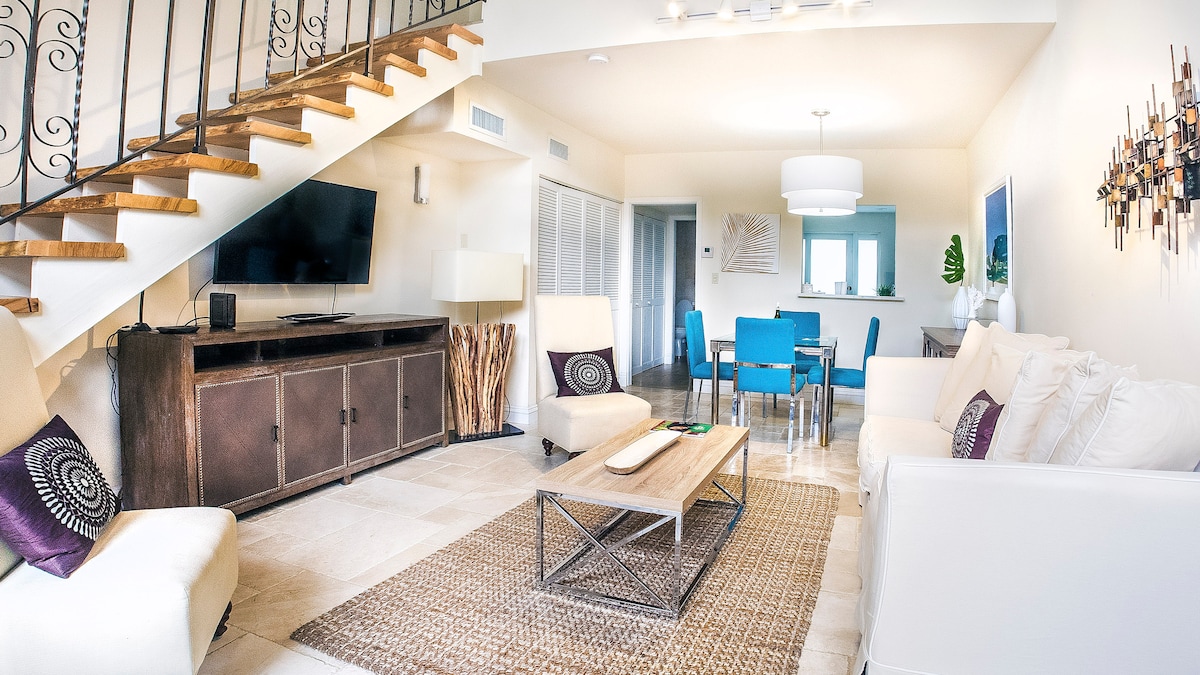 An open living area features plush sofas and a coffee table on a woven rug. A large TV is mounted on the wall opposite a decorative storage unit. Natural light fills the space, highlighting the dining area with a blue table set for four. A staircase leads to the upper level.