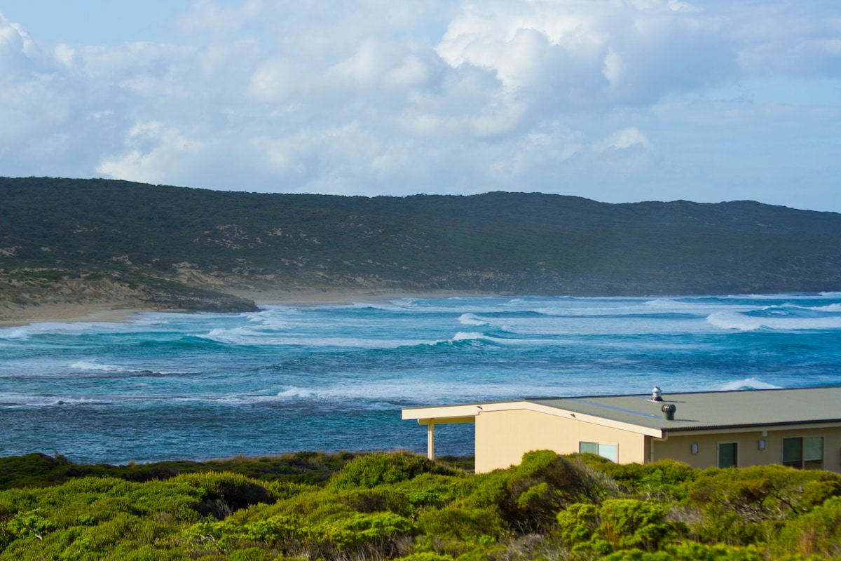 The image captures a view of the ocean waves rolling onto a rugged coastline, with lush greenery visible in the foreground. The gentle curves of the hills in the background contrast with the expansive blue waters, creating a serene natural landscape.