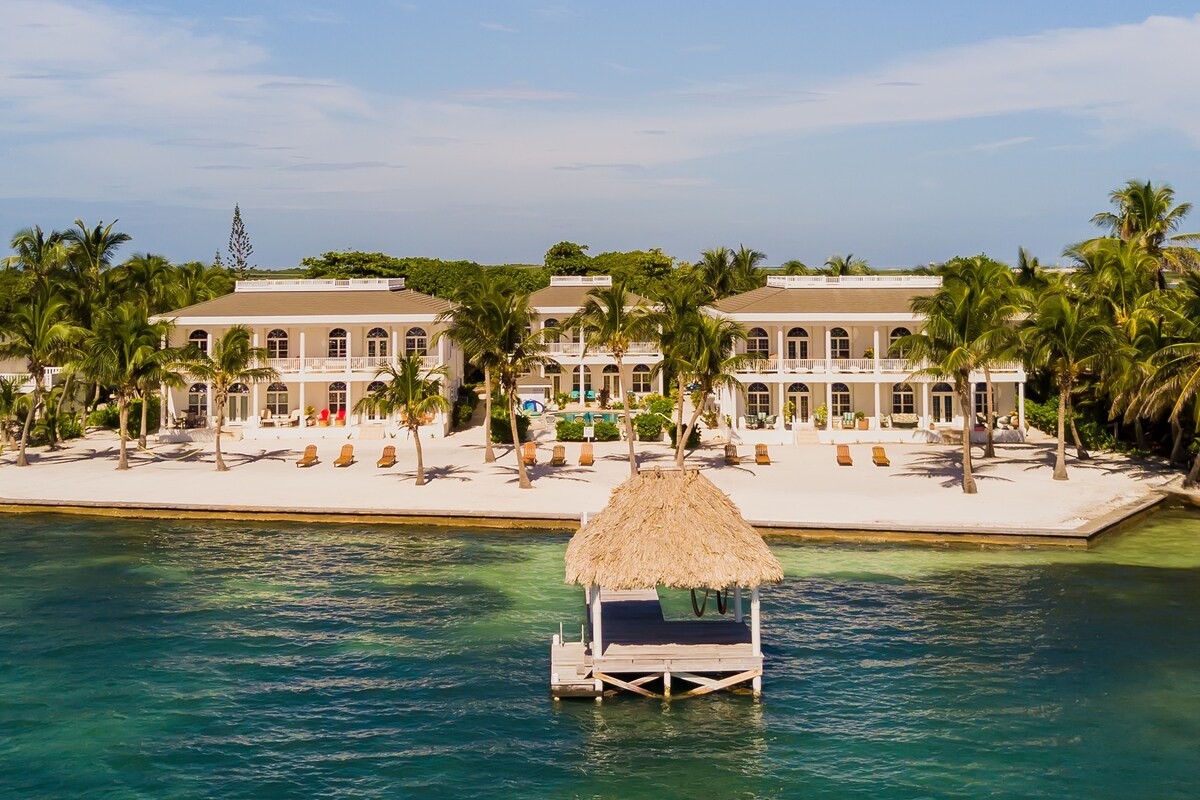 The beachfront resort is surrounded by lush palm trees, with sandy areas for lounging. A charming palapa stands on the water, offering a shaded spot for relaxation. Several lounge chairs are arranged on the beach, inviting guests to enjoy the serene atmosphere and views of the Caribbean Sea.