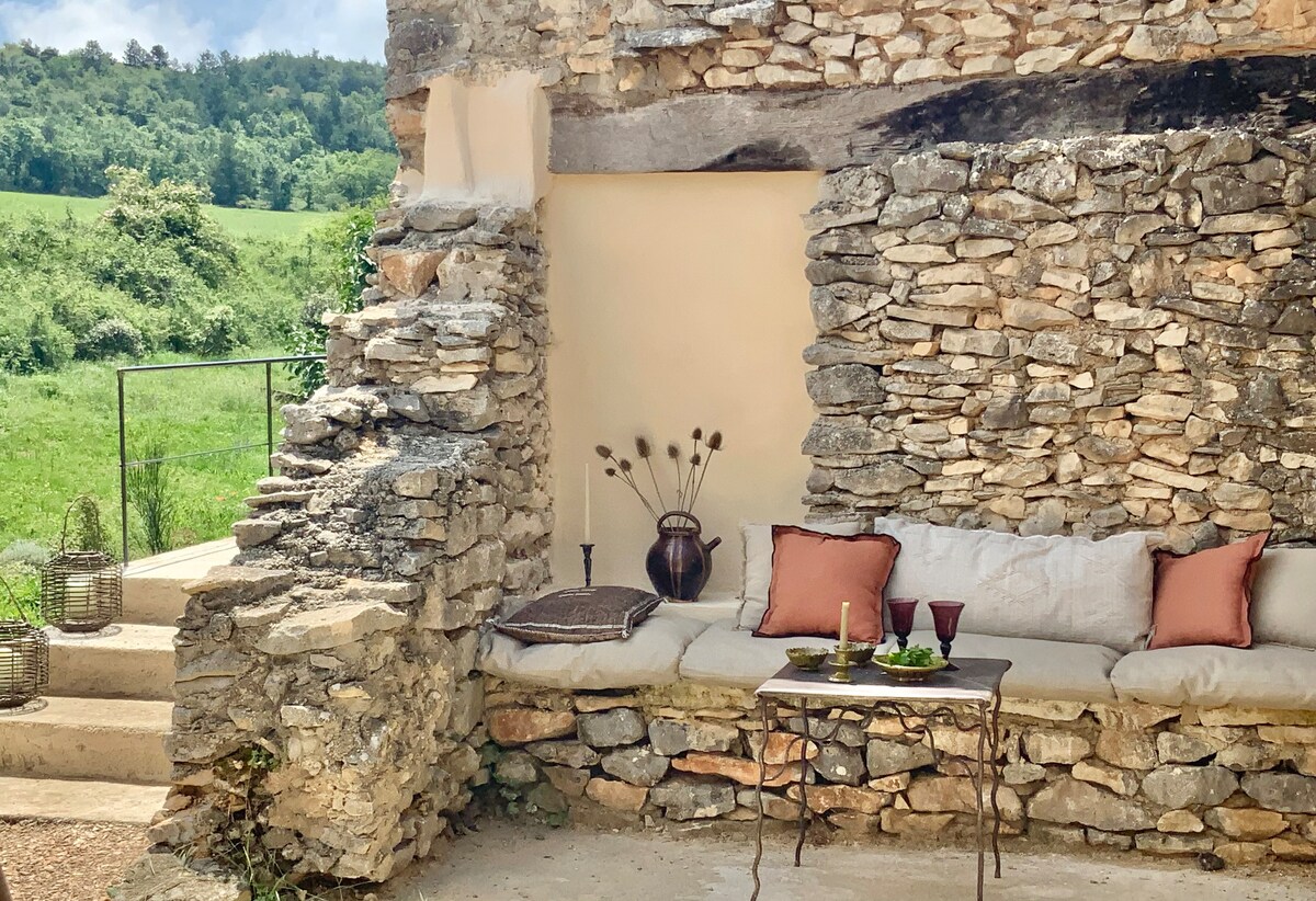 A charming outdoor seating area is framed by natural stone walls, providing a rustic yet comfortable space. A table is set with candles and decorative items, while plush cushions add a touch of comfort. Lush greenery is visible in the background.