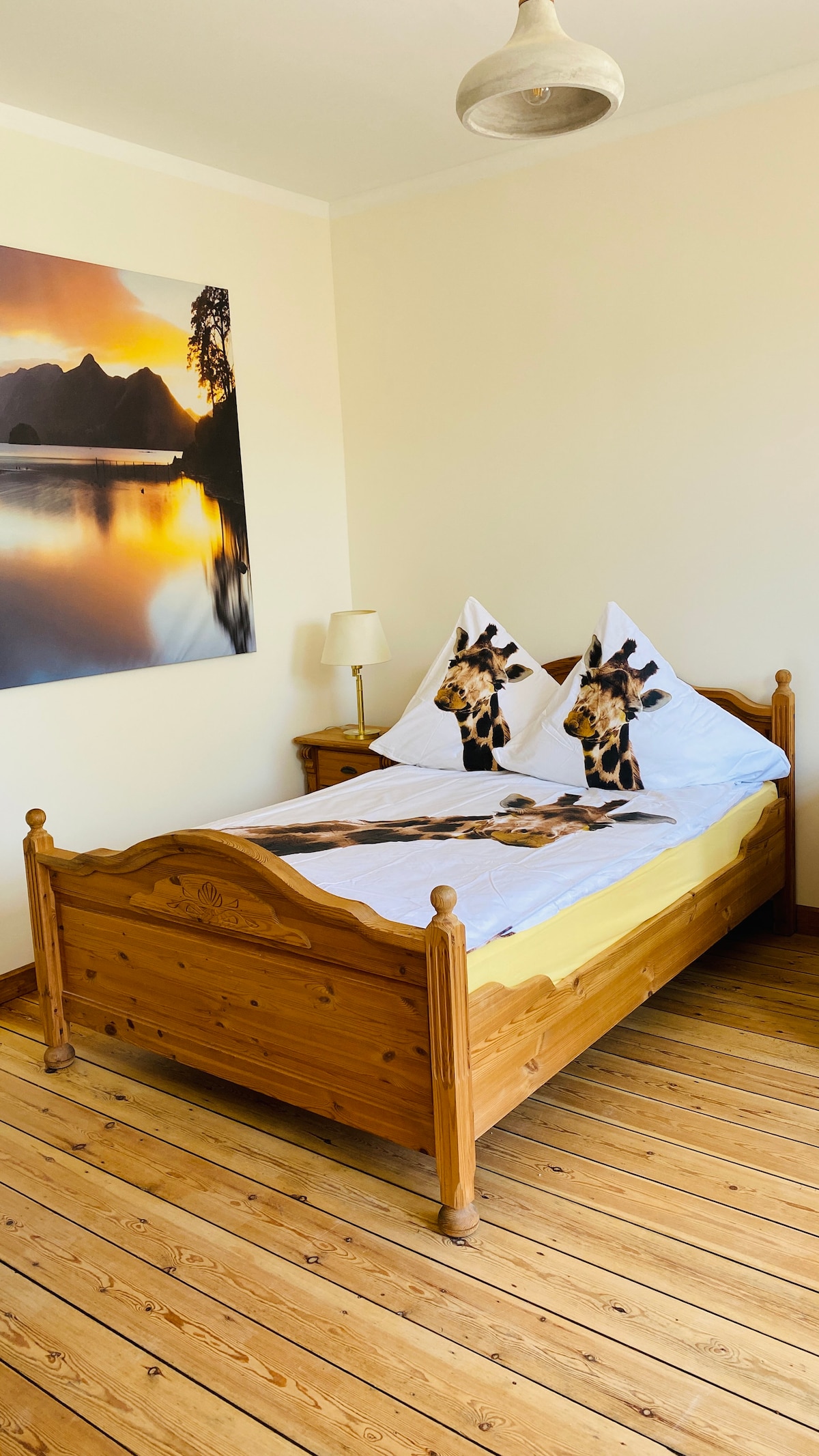 A wooden bed with a light yellow blanket is positioned against a wall. Two decorative pillows featuring a giraffe design are arranged on the bed. A bedside lamp is on a small table, and a scenic landscape photograph hangs on the wall, complementing the natural wood flooring.