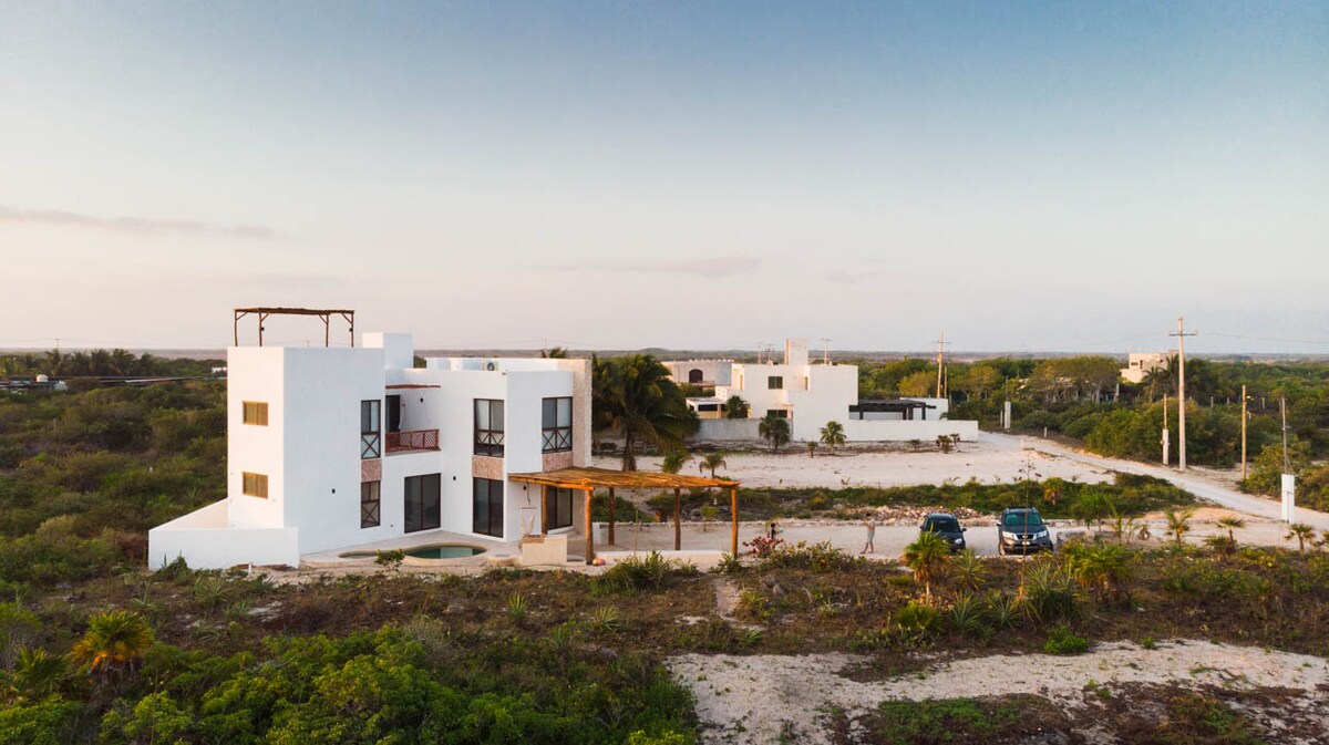 An aerial view of a modern white house surrounded by natural vegetation. The property features a spacious outdoor area with a wooden pergola and parking spaces. Nearby structures are visible in the distance against a clear sky.