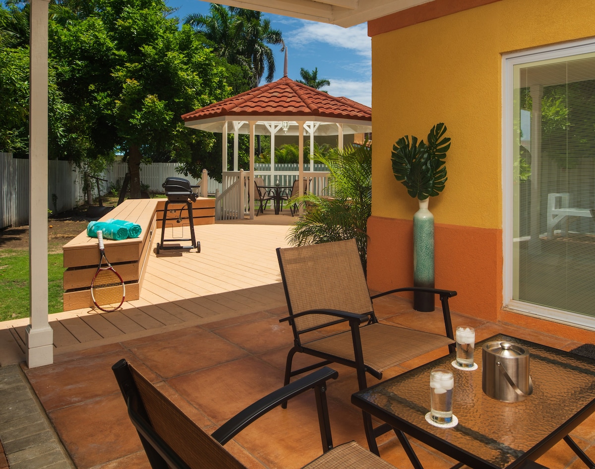 A cozy outdoor patio features a glass-top table with two chairs, a decorative vase, and a small set of candles. A grilling area is visible in the background, alongside a gazebo and a patio space adorned with lush greenery.