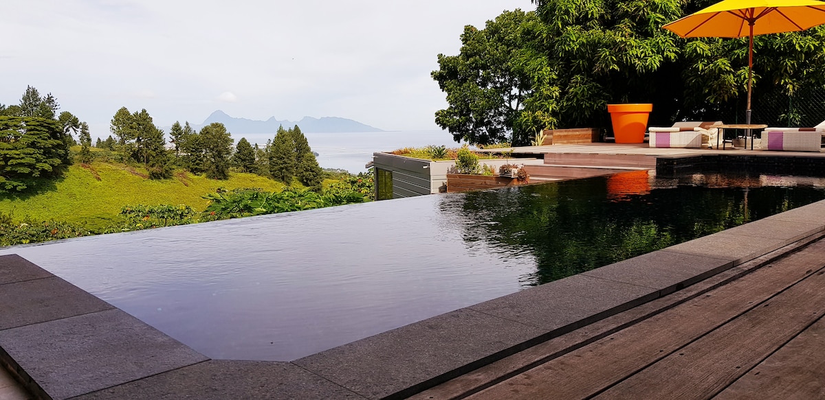 An infinity pool mirrors the serene landscape, with lush green foliage surrounding the area. A bright orange umbrella provides shade over a seating arrangement, while distant mountains are visible across the water. The pool's edge creates a seamless transition to nature.