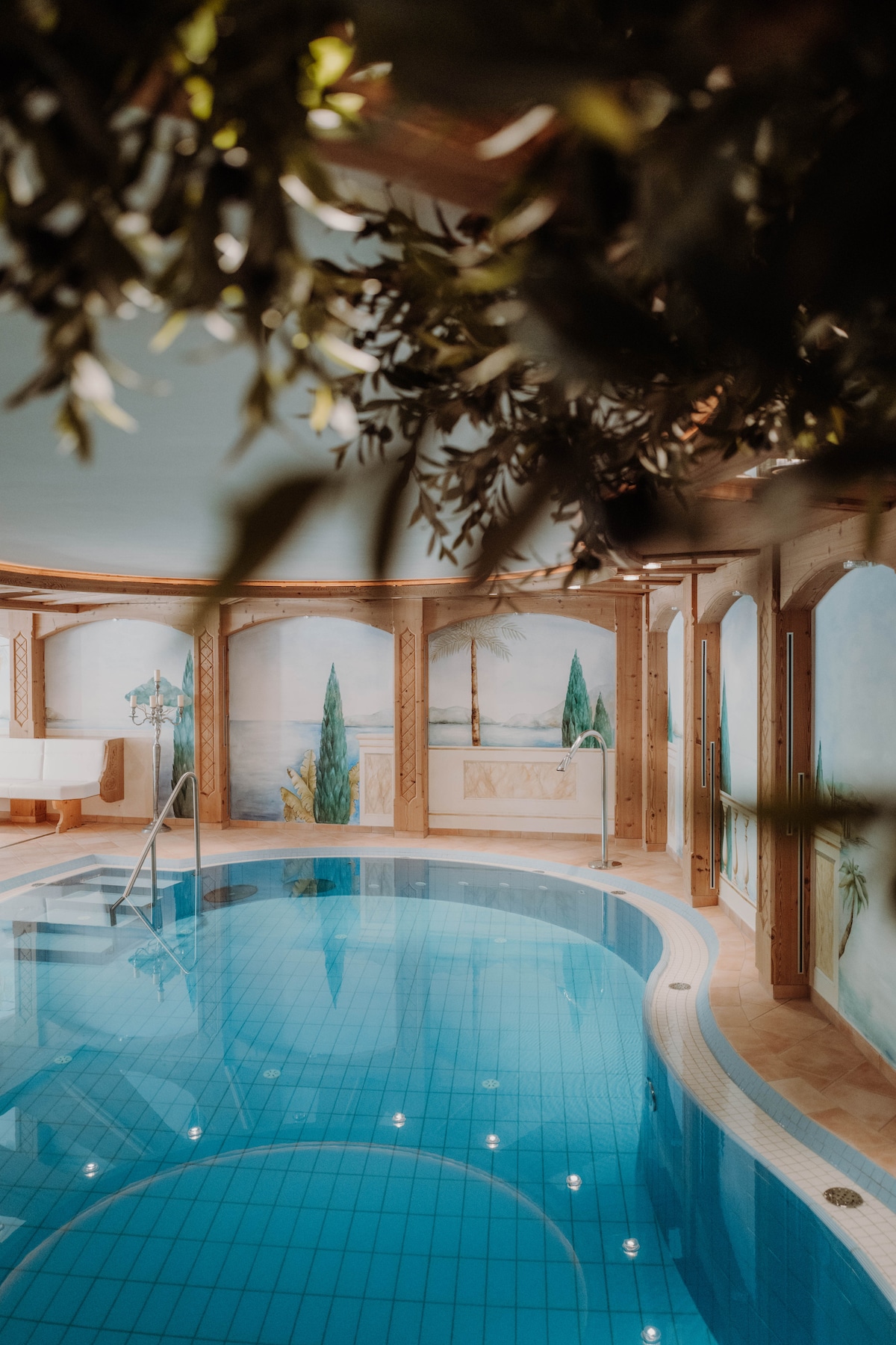 An indoor pool area features crystal clear water with subtle blue tiles. Surrounding walls are adorned with scenic murals depicting cypress trees and coastal views. Soft lighting reflects off the water, creating a serene ambiance. A staircase provides entry to the pool area.