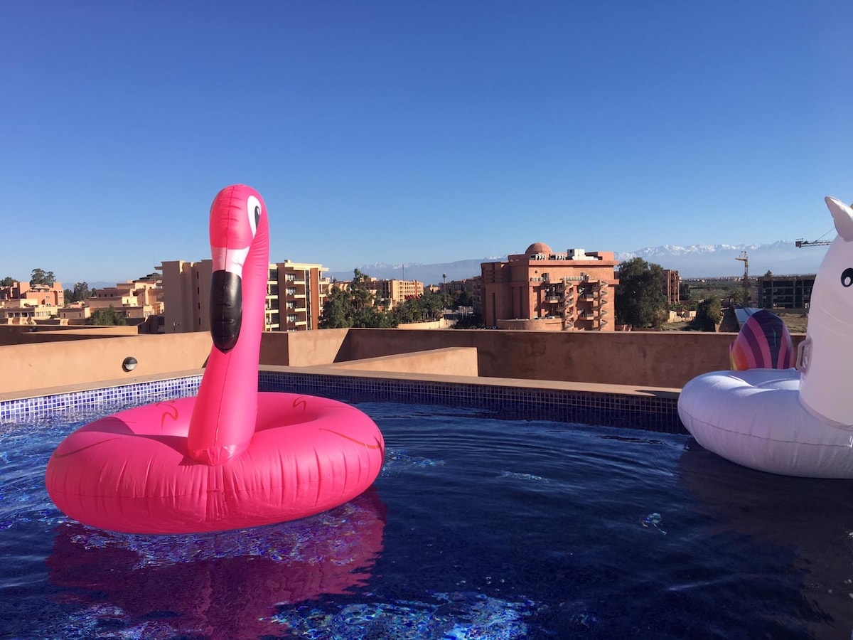 A rooftop pool features playful inflatable decorations, including a pink flamingo and a unicorn. The surrounding area offers clear blue skies and distant views of the Atlas Mountains, with pool tiles reflecting sunlight on the water's surface.