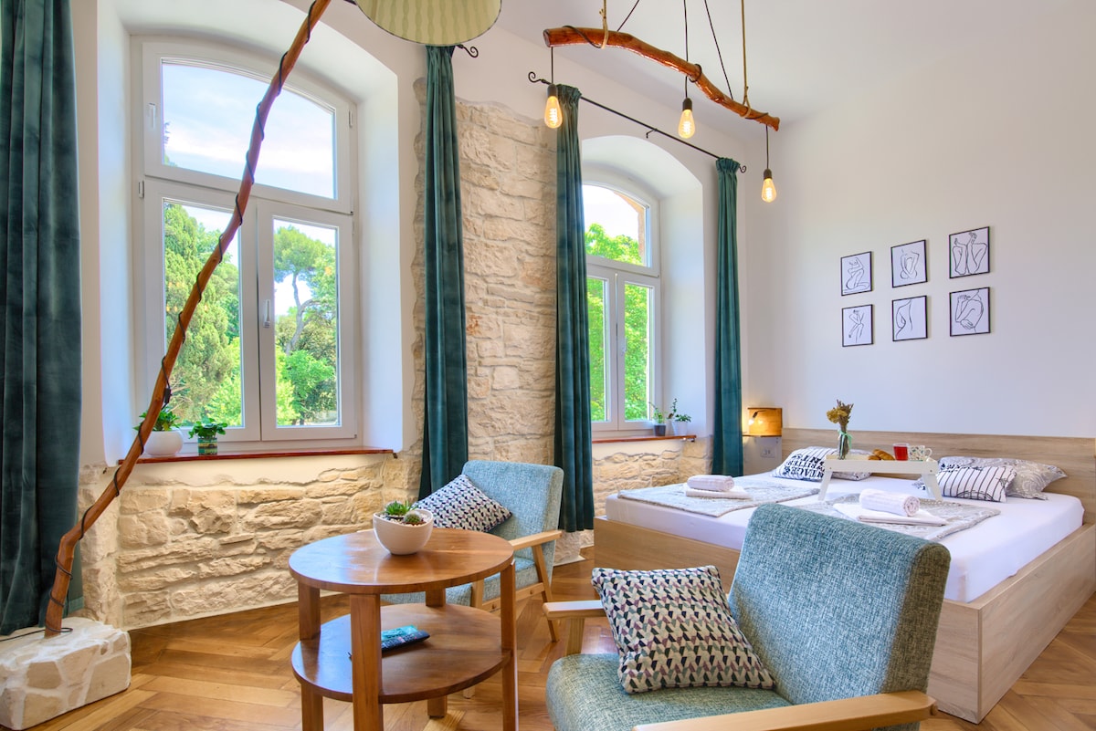 A modern and bright room features large windows that provide a view of the surrounding greenery. Light wood furniture includes a spacious bed and a small dining table with two chairs. Textured walls and soft curtains contribute to a serene ambiance.