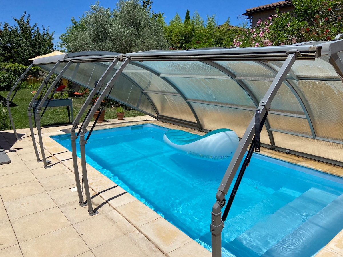 A covered swimming pool is displayed, featuring clear water reflecting sunlight. The pool is enclosed by a transparent structure that offers protection while allowing visibility. Surrounding greenery is visible, with manicured plants enhancing the outdoor space.